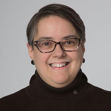 A smiling woman with graying dark hair and glasses, wearing a black turtleneck and black earrings