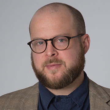 A man with close cropped dark hair, a beard, and glasses, wearing a dark shirt and brown jacket