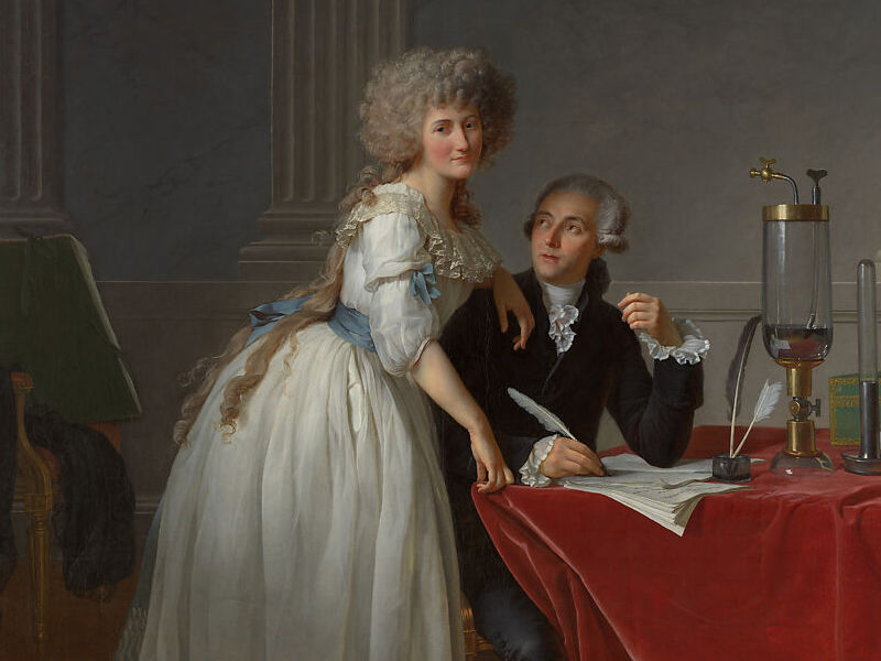 Portrait of Antoine Laurent Lavoisier and Marie Anne Lavoisier. An 18th century French couple wearing white wigs. Marie is standing and wearing a white dress. Antonie is sitting, wearing a black suit, and writing with a quill pen on documents sitting on a table draped with a red table cloth and covered with scientific instruments.