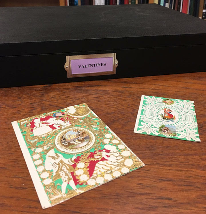 Photo of a black box labeled 'VALENTINES' on a wooden table with two historic valentines laid out on the table top