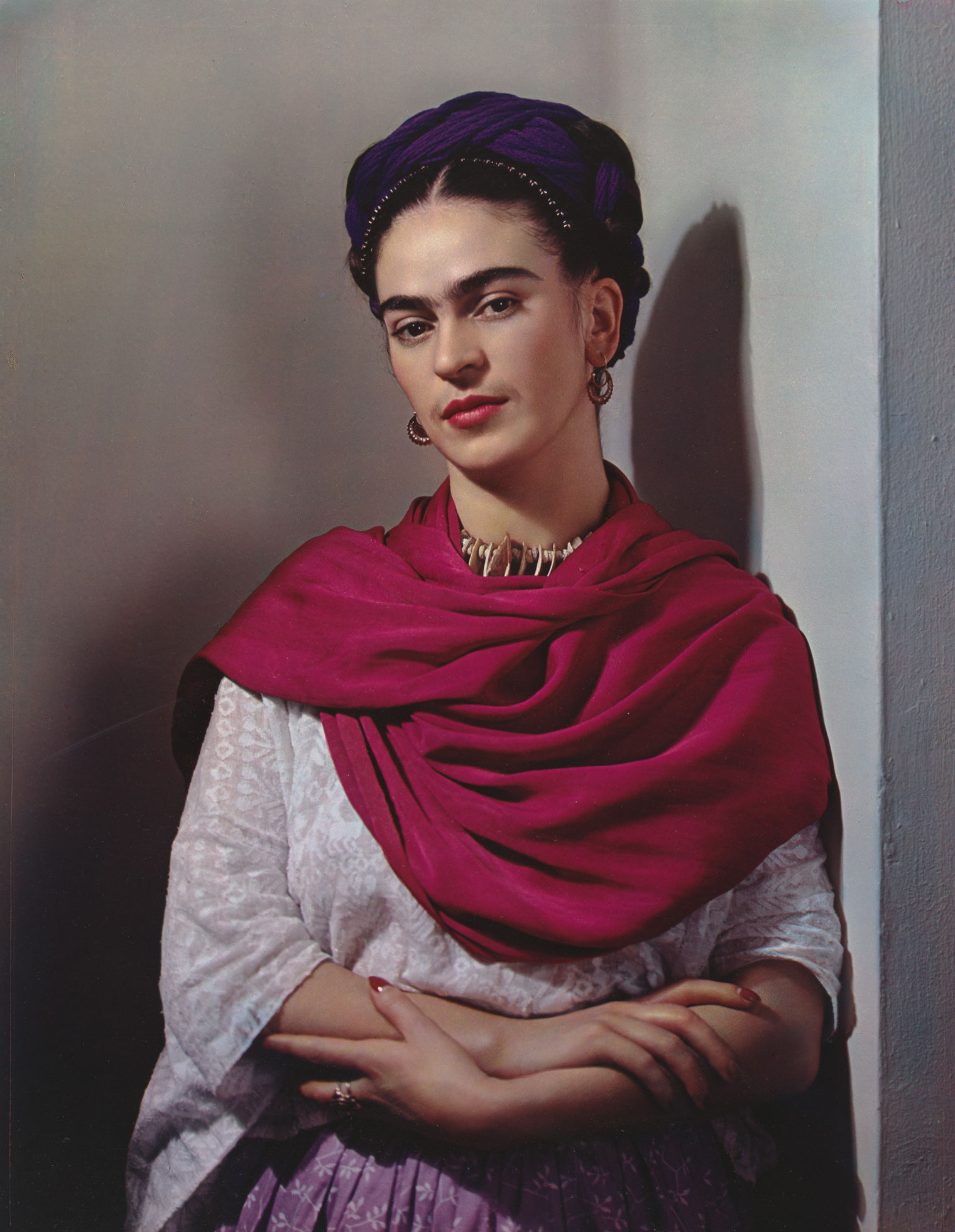 Photo portrait of Frida Kahlo, leaning against a wall with her forearms crossed as she gazes at the camera