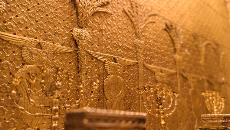 An ornate gold color wall in the interior of a miniature temple, with menorahs on two tables against the wall.