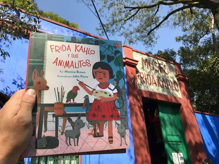 A hand holds a copy of the book "Frida Kahlo and her Animalitos" in front of the entrance of "the blue house" La Casa Azul. Over the red doorway of the building read the words "Museo Frida Kahlo" in tile.