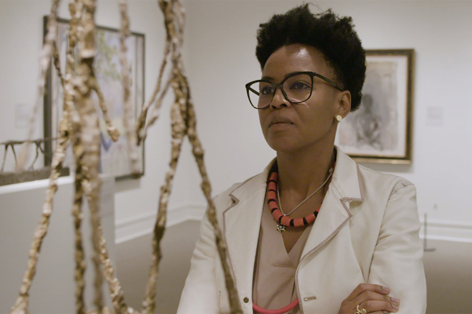 The artist Wangechi Mutu, a tall woman in a light coat and glasses, looks at a Giacometti sculpture of three elongated humans