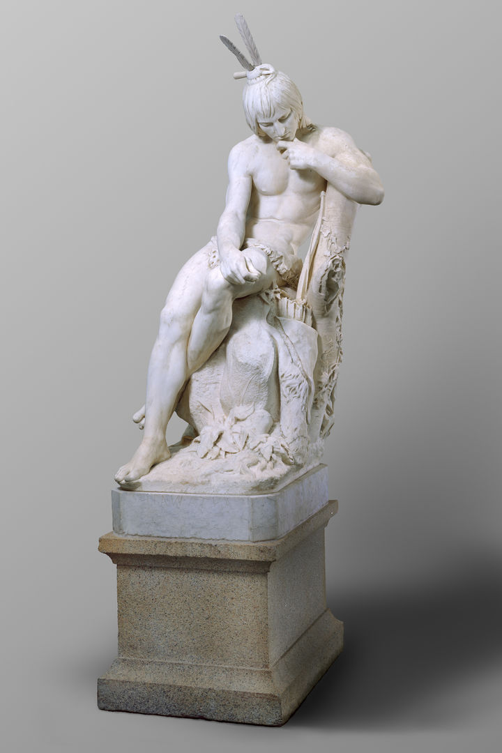 A white marble sculpture of an Indigenous man, seated in contemplation