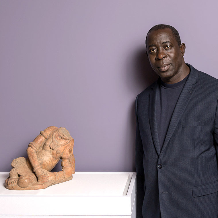 A tall man with dark skin, wearing a navy blue suit, stands next to a terracotta figure of a nude woman, in front of a purple background