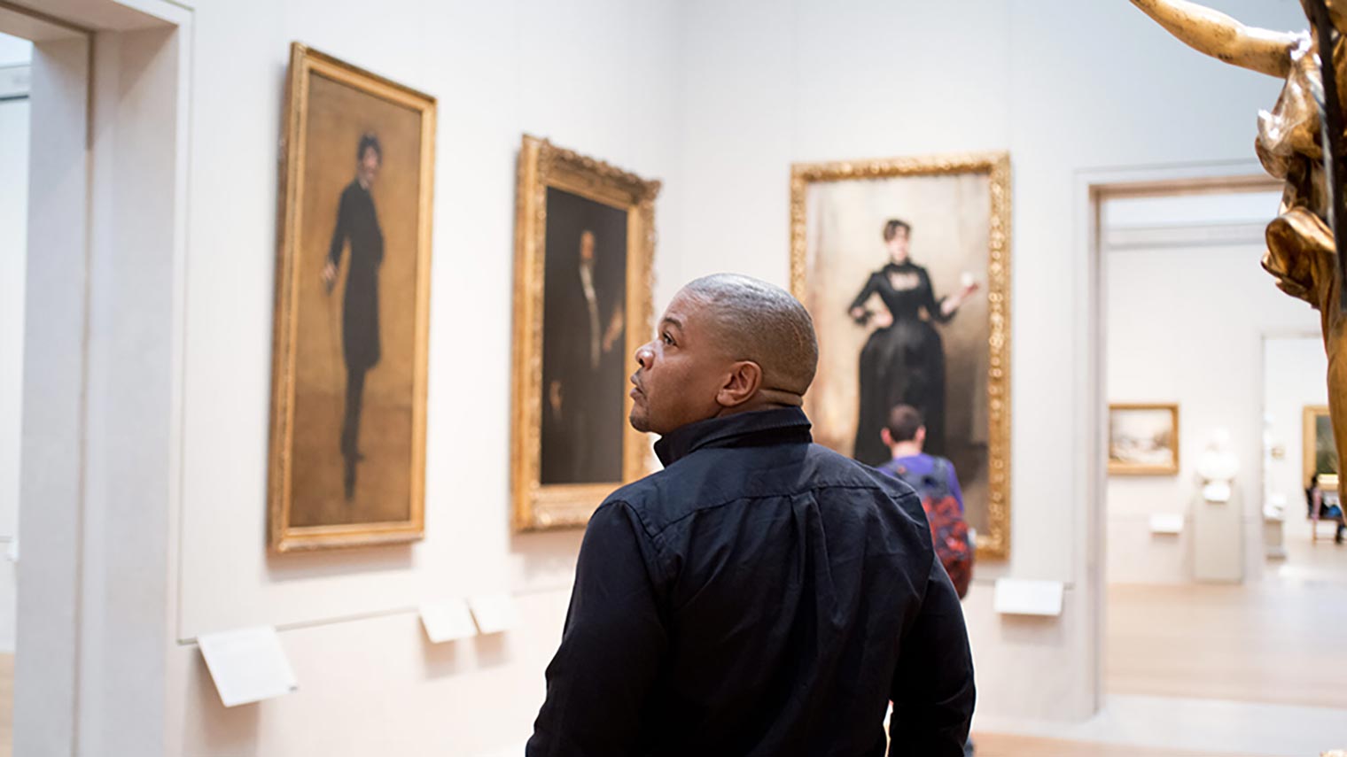 A photograph of the artist Kehinde Wiley walking through the Met's American Wing