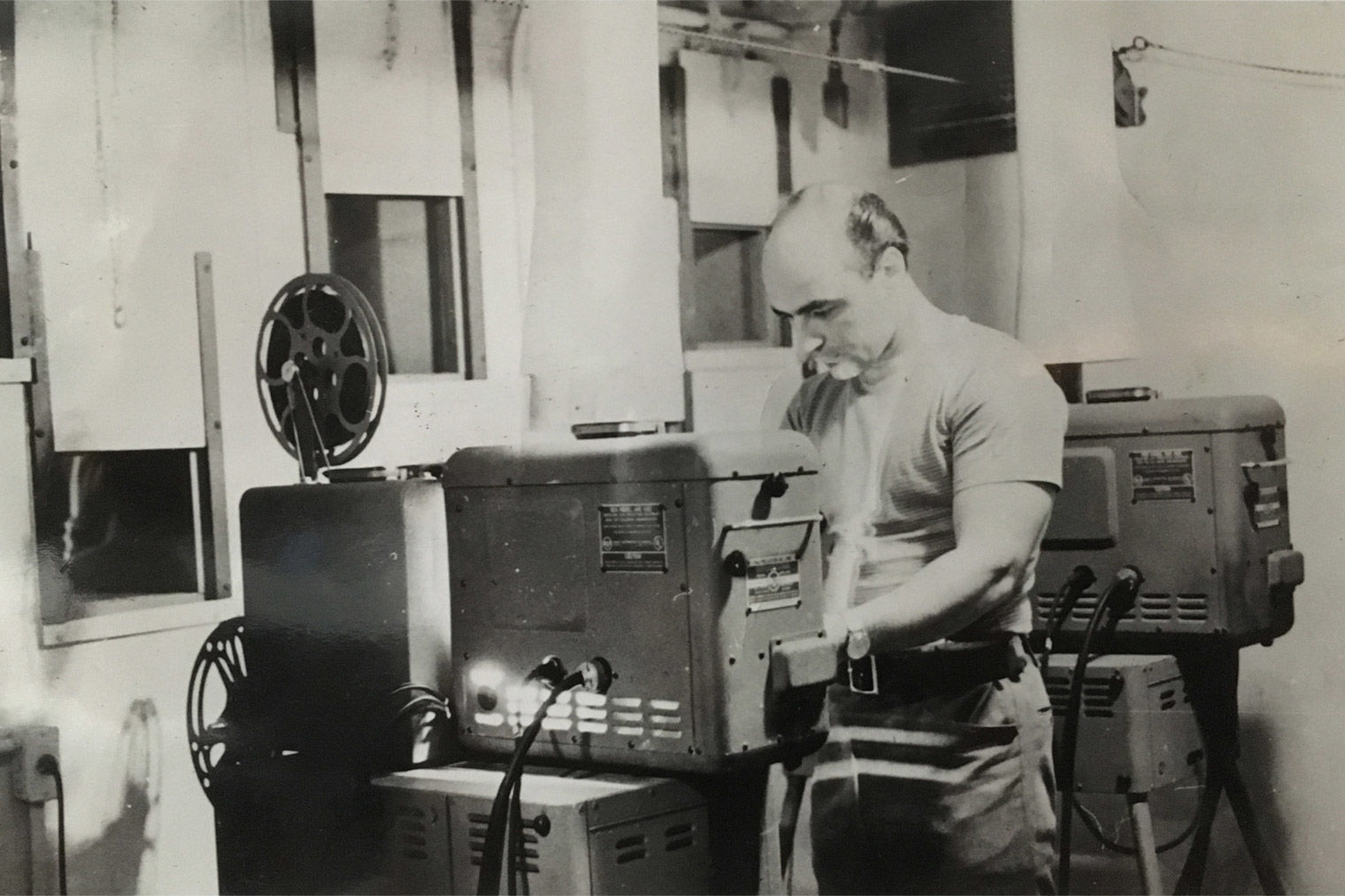 A black-and-white photo of the artist and audio-visual technician Ray Cusie trimming the carbon arc of a 16-mm projector in the Junior Museum auditorium booth.