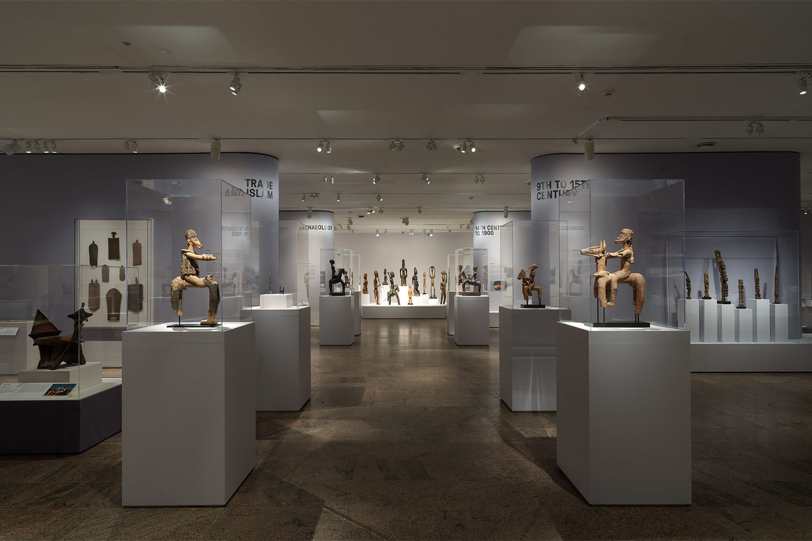 Two rows of wooden figurines on display stretch across the room into a vanishing point of a white platform showcasing similar objects.