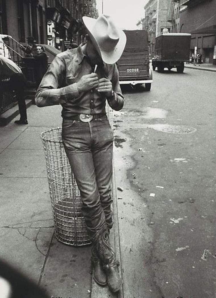 Black and white photograph of a man with a cowboy hat smoking as he leans against a trash bin