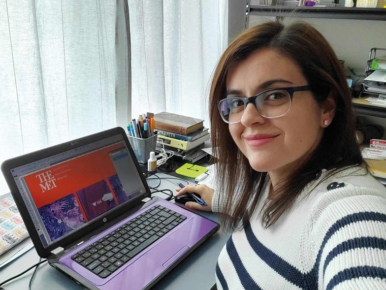 Selfie of a smiling young woman seated at a desk in an apartment, in front of a laptop with The Met's website displayed onscreen.
