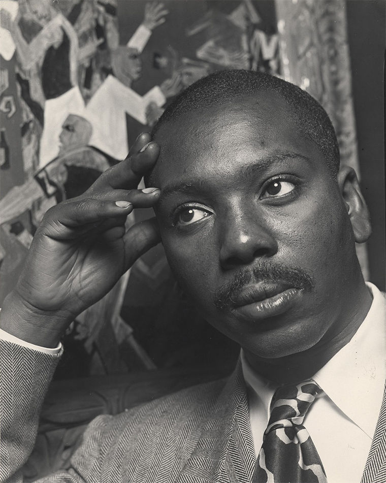 A black-and-white photo of Jacob Lawrence resting his head in his hand in front of one of his paintings