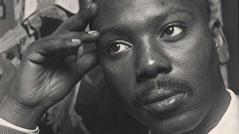 A black-and-white photo of Jacob Lawrence resting his head in his hand in front of one of his paintings