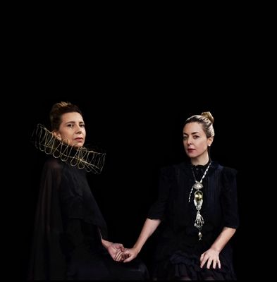 Two women, one wearing a wire outline of a ruffled collar, hold hands and look into the camera.