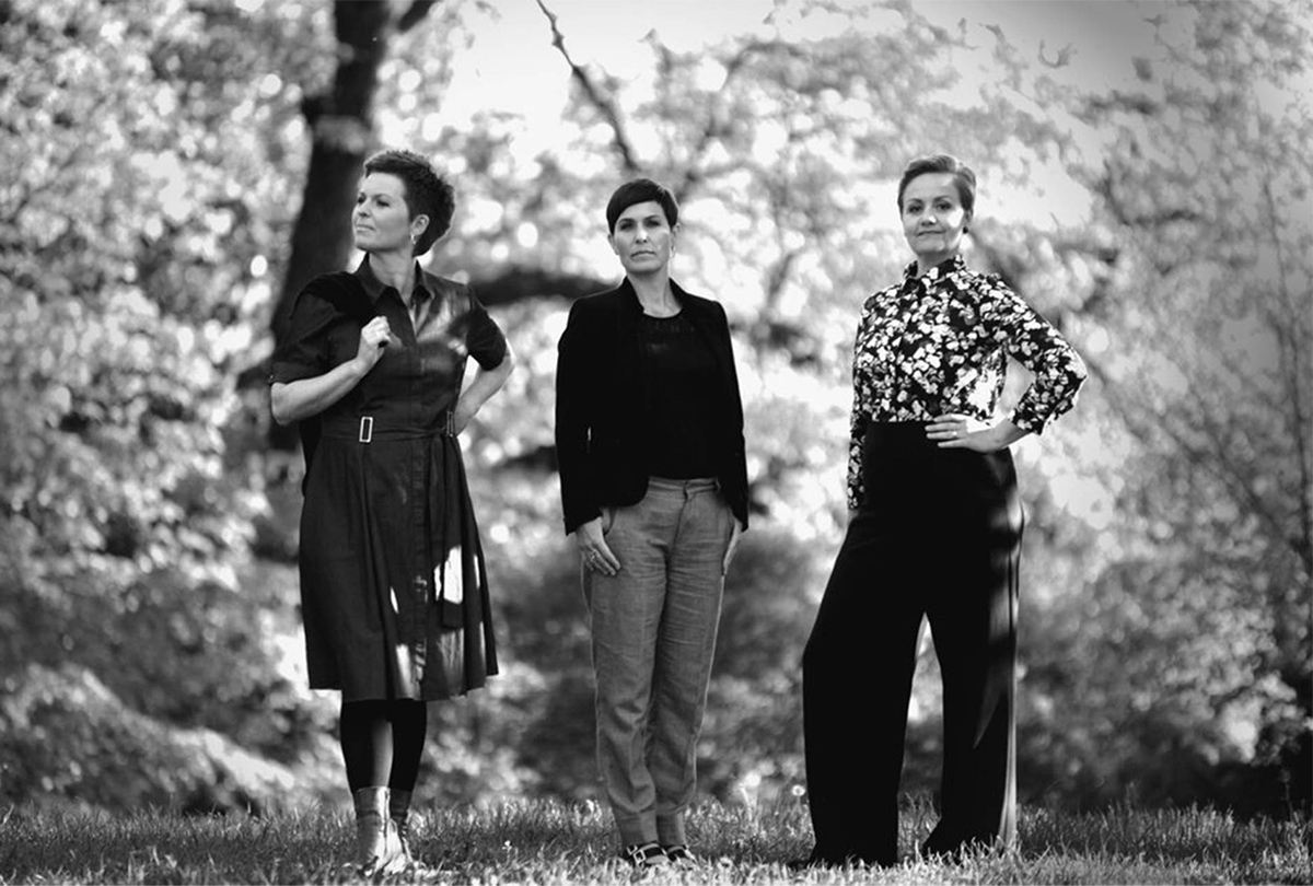 A black and white image of three women posing outside amid trees.