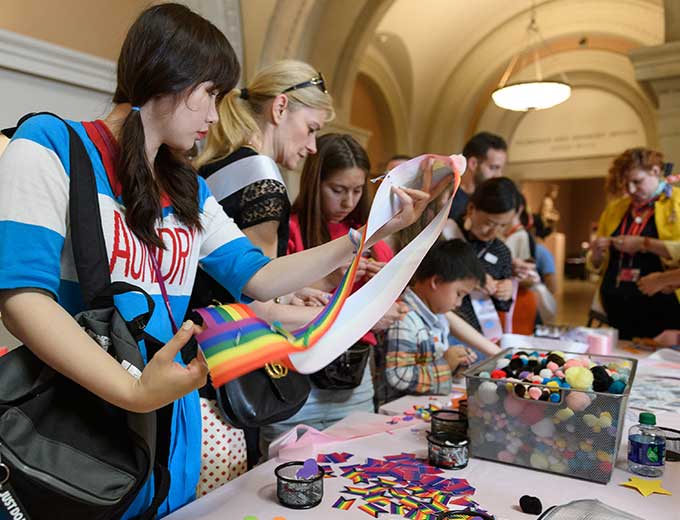 Young people are standing at a long table and selecting arts and crafts materials such as colored paper and fabrics.