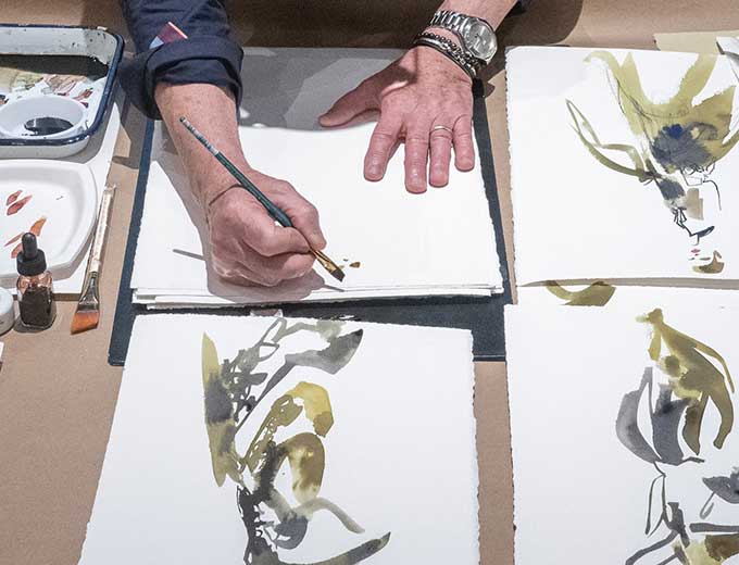 A man stands at a table painting abstract figures on white paper.