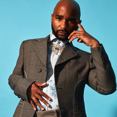A headshot of a bald man in a zip-up collared shirt, a grey tweed jacket, and a pendant around his neck. One hand is rested against his head. The other is on his stomach.