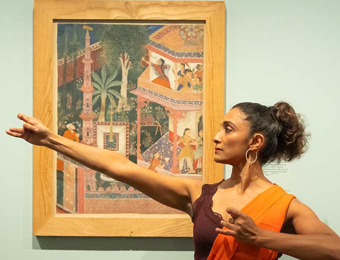 An Indian woman with hair tied into a ponytail and wearing a burgundy camisole top and a bright orange and yellow scarf wrapped across her torso, is seen from her left side extending her right arm as her left elbow pulls back in space. She stands in front of a colorful sixteenth century Mughal painting that depicts the archeress Mihrdukht shooting an arrow amid onlookers in a court garden.
