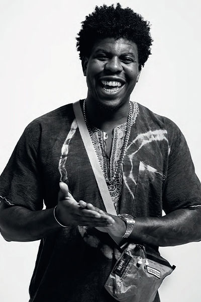 Black-and-white photo-portrait of Jon Gray. He has a wide, joyful smile. His hands are in front of him as though he is applauding. He is wearing an open neck shirt and a cross-body bag.