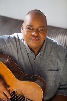 Toshi Reagon wears a blue button-down shirt. Her hair is closely shaven, and she wears a hooped earring in her right ear. She looks relaxed and directly at the camera. Her right hand rests on the strings of her acoustic guitar as if she is about to play.