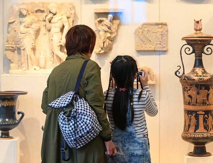 Mother and daughter standing with backs turned in front of Greek & Roman display case.