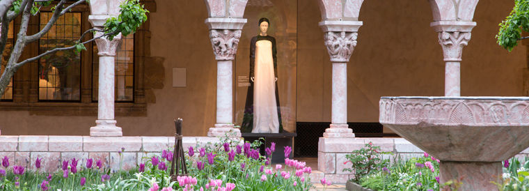 Gallery view of "Heavenly Bodies" showing contemporary fashions inspired by medieval art juxtaposed with medieval art objects in a cloistered garden