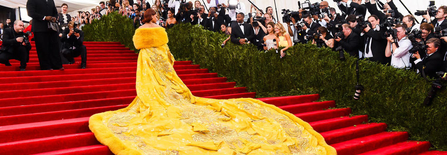 Rihanna walks up the red carpet steps of The Met Fifth Avenue wearing a dramatic yellow gown by Guo Pei and flanked by photographers