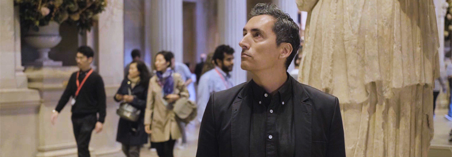 The artist Kent Monkman, wearing a black suit and jacket, stands in the Neoclassical Great Hall