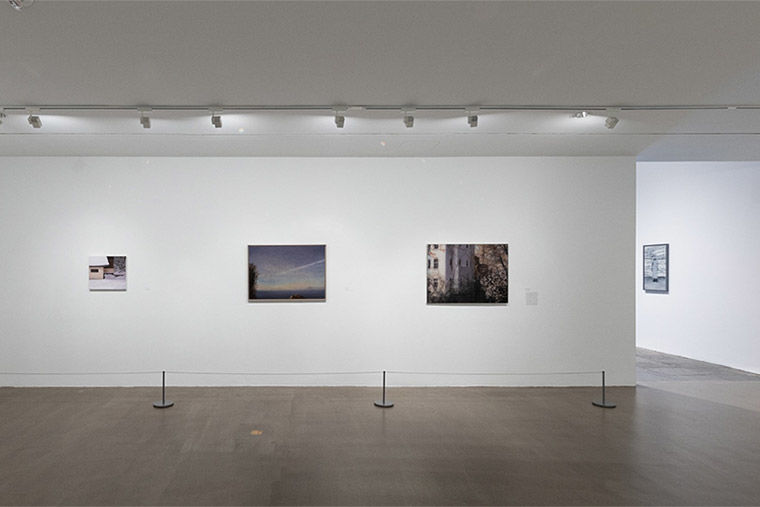 Installation view of Gerhard Richter: Painting After All at The Met Breuer featuring three paintings; a house in snow, an open sky at dusk, and a building overgrown with foliage