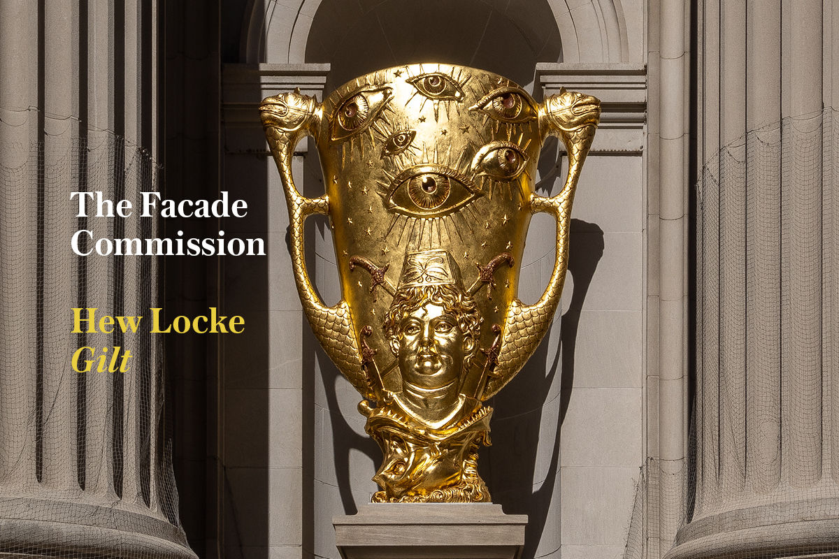 The Facade Commission: Hew Locke, Gilt text next to one of his shining gold sculptures in The Met's niches. It is a large gilt goblet dotted with starry eyes, a central cherubic male figure, sabers, and a serpentine monster devouring all at its base.