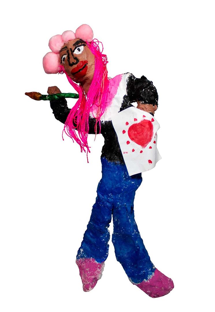 Sculpture of a smiling dark-skinned person with pink hair, wearing a dark top, blue pants and pink shoes. The person is holding a white card with a red heart.