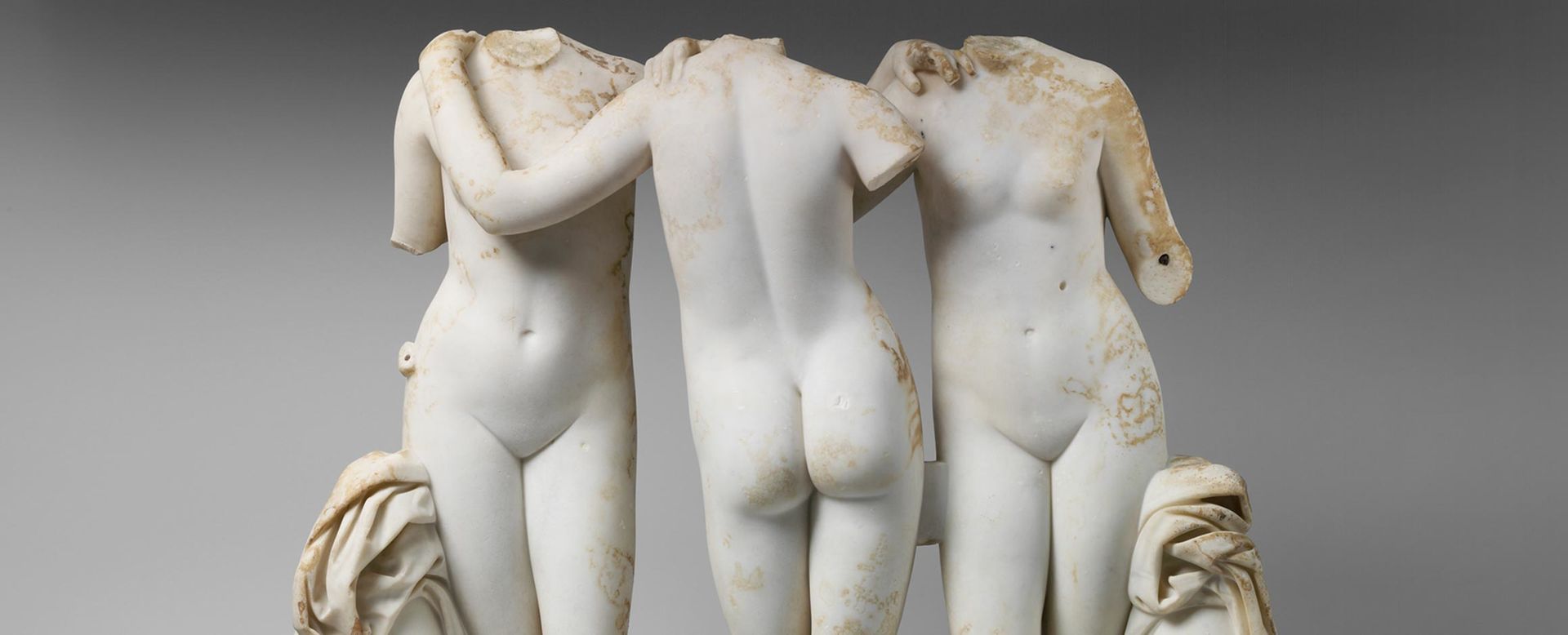 Marble statue of three young women standing in contrapposto between two urns, with fabric on top, while holding on to each other's shoulders