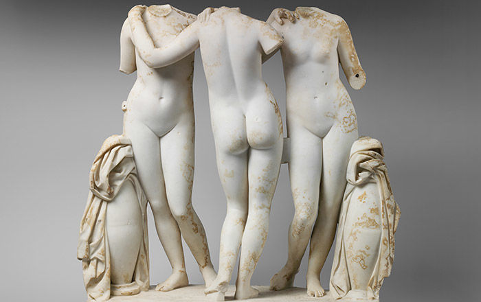 Marble statue of three young women standing in contrapposto between two urns, with fabric on top, while holding on to each other's shoulders