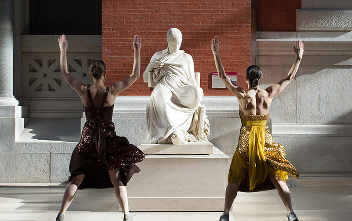 Two standing women, facing a statue of Sappho on opposites sides, with arms and legs spread during a museum workout