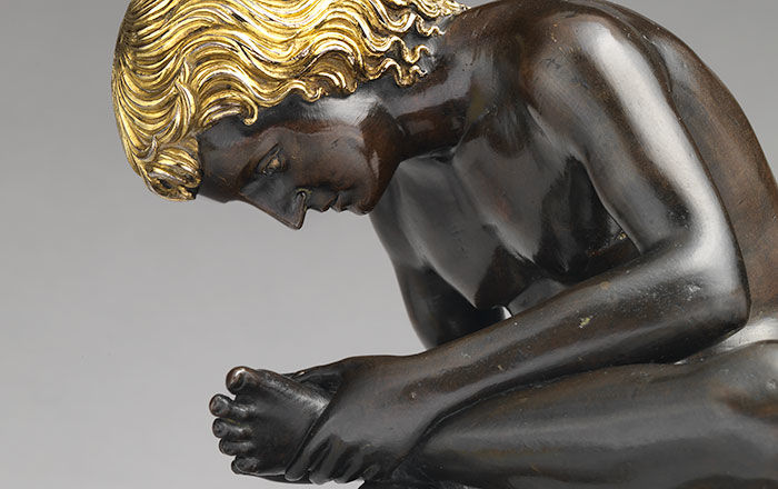 Bronze sculpture of a young boy with gold wavy hair pulling out a thorn from his foot while sitting on a tree stump
