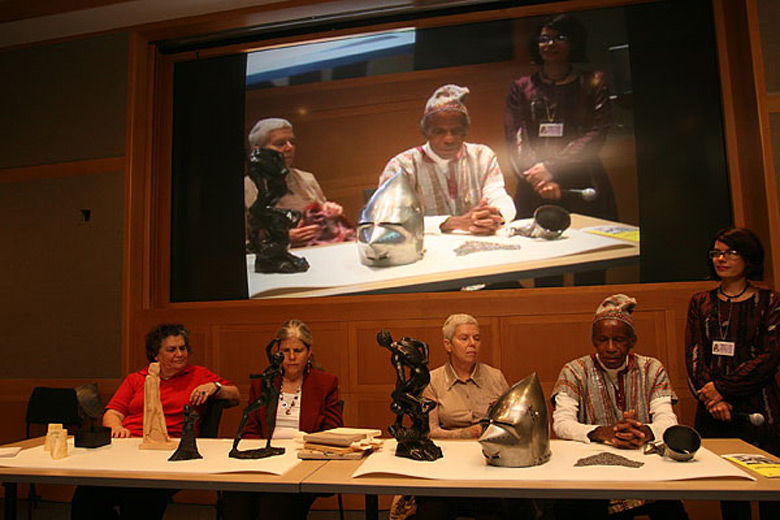 Five people at a table with sculptures and art objects on it; behind them is a large screen with a projection of the live procedings