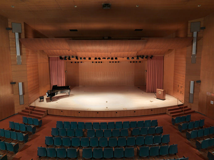 An elevated view of a large modern blonde wood auditorium as viewed from the balcony; in the foreground are blue auditorium seats in three sections