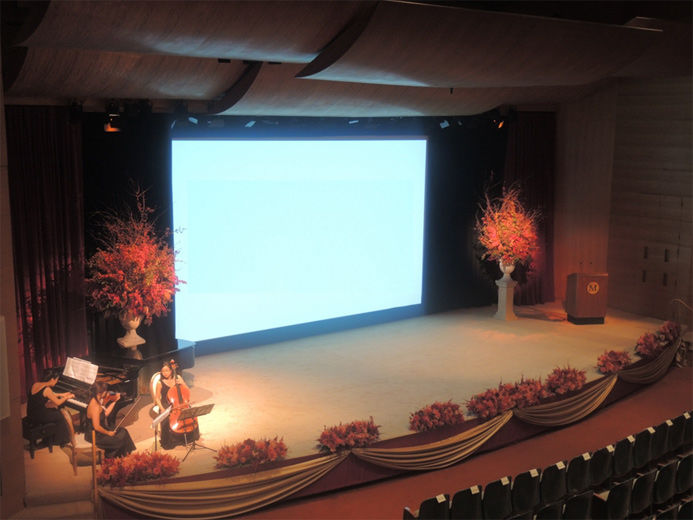 An elevated view of a large modern blonde wood auditorium as viewed from the balcony; on stage, the screen is lowered to show a projection and on either side of the screen are large vases filled with branches of autumn leaves red berries; stage right is a female classical musical group playing the piano, violin and cello