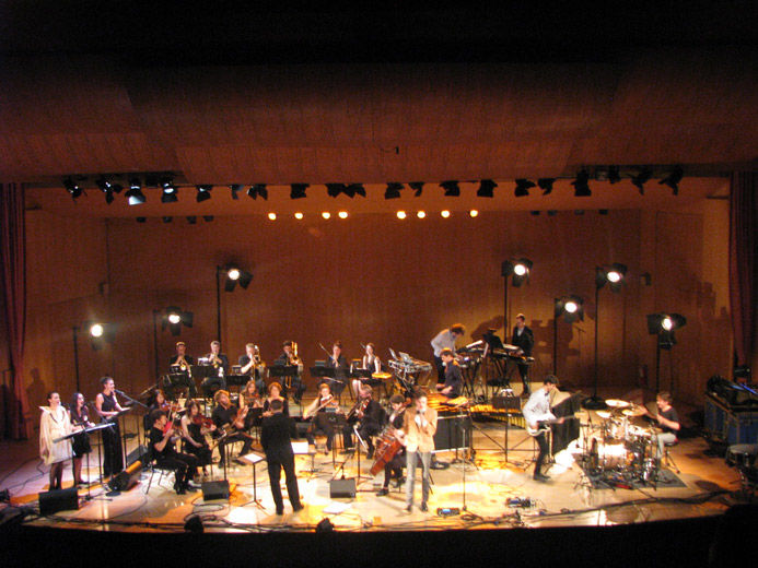 An elevated close-up of the stage of a large modern blonde wood auditorium as viewed from the balcony; on stage, is a large, modern orchestra