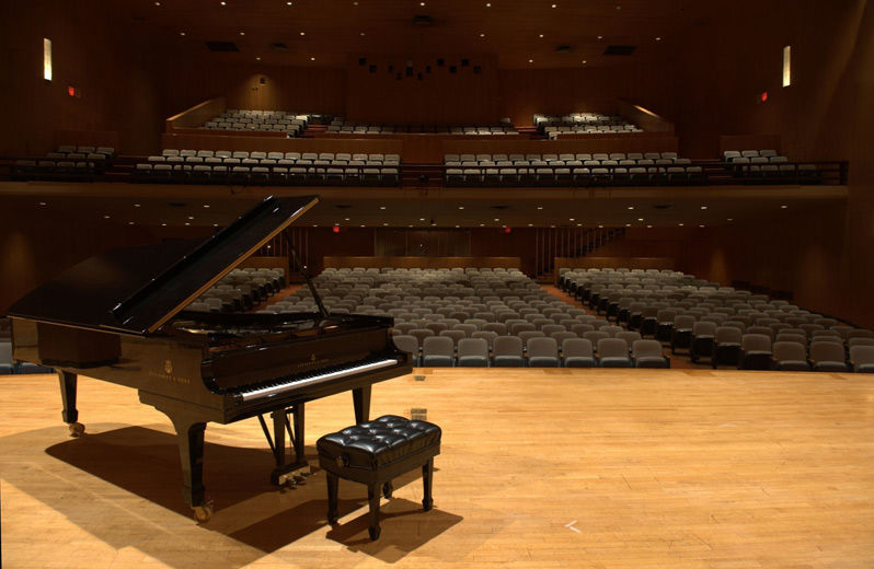 A large modern auditorium with two-tiered balcony as viewed from the stage; on the left side of the stage is a black grand piano with the lid and fallborad raised, and a padded piano bench in front