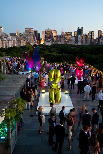 An elevated view of a rooftop sculpture garden with skyline views, at dusk; on display are large reflective sculptures in the shape of a balloon-animal-dog, and a heart