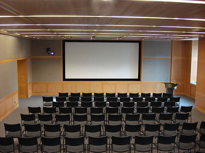 A large, comfortable, carpeted, modern, lecture hall with blonde wood and grey fabric paneling; the room is set with rows of chairs facing a large projection screen