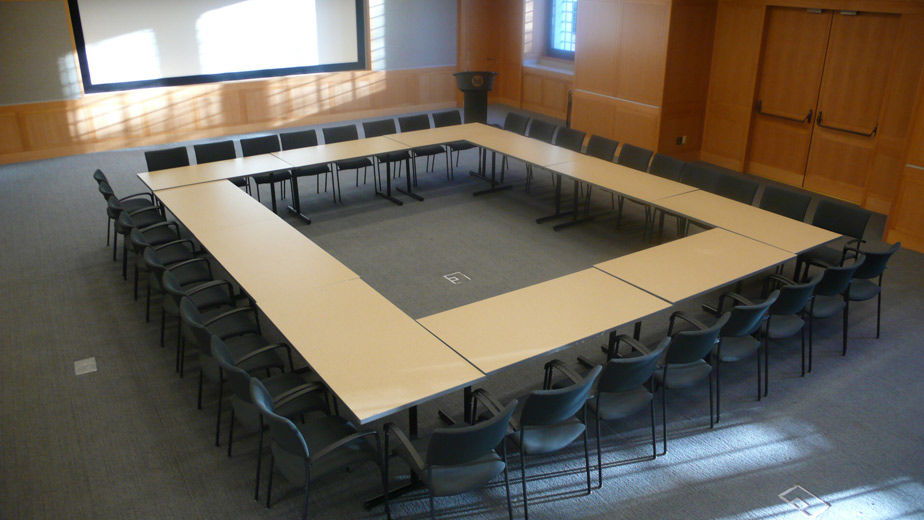 A large, comfortable, carpeted, modern, lecture hall with blonde wood and grey fabric paneling; the room is set with long tables in an enclosed rectangular shape and chairs on the perimeter