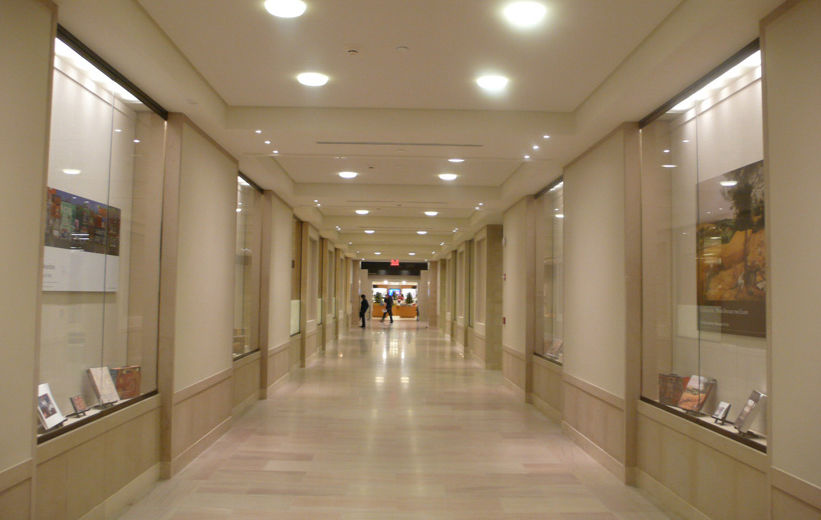A long, brightly lit corridor with white stone flooring and light beige fabric walls; inset along the walls are tall, shallow glass display cases 