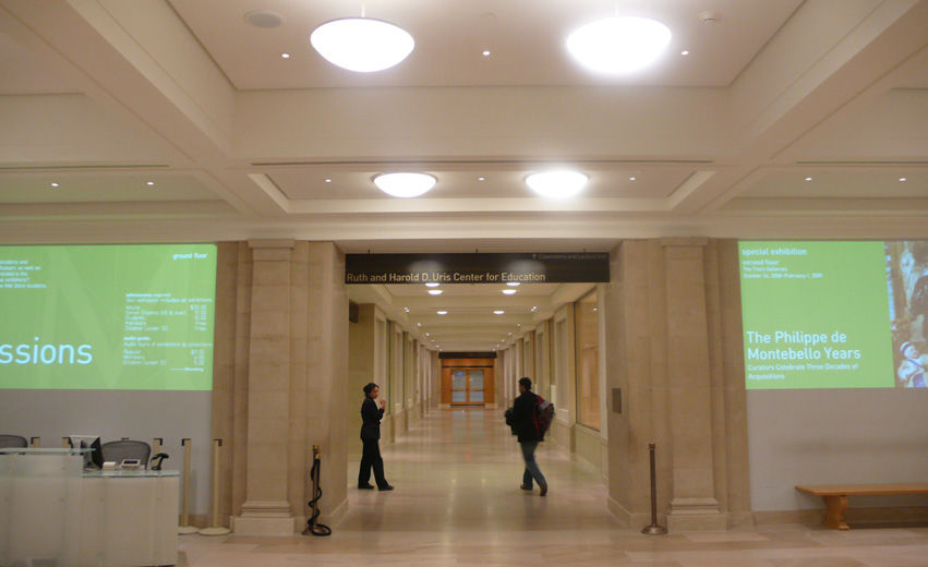The entryway to long, brightly lit corridor with white stone flooring and light beige fabric walls; on the walls on either side of the entryway are projections of white text on a green background