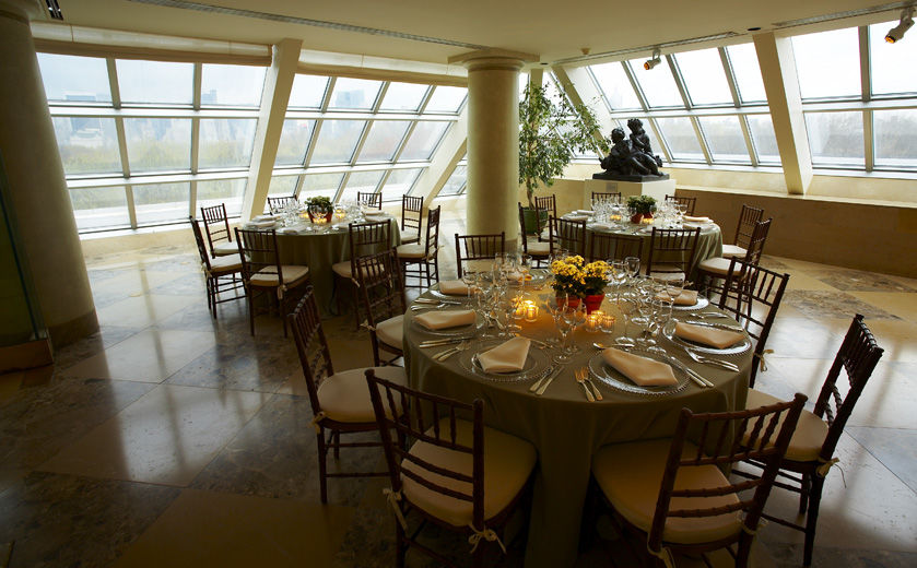 A sunlit interior terrace set with round dining tables draped in light green table cloths and formal table service