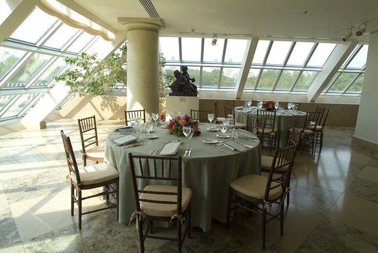 A bright, sunlit interior terrace set with round dining tables draped in light green table cloths, casual table service and colorful, large, low flower arrangements