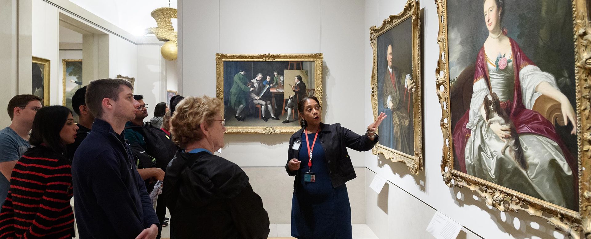 A female volunteer leads a group of visitors on a tour in the American Wing. She is gesturing towards and discussing an oil painting in The Met collection.