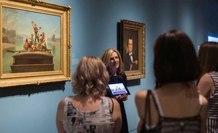 Visitors are listening to a tour guide who is standing in front of an oil painting, holding an iPad in a blue gallery.